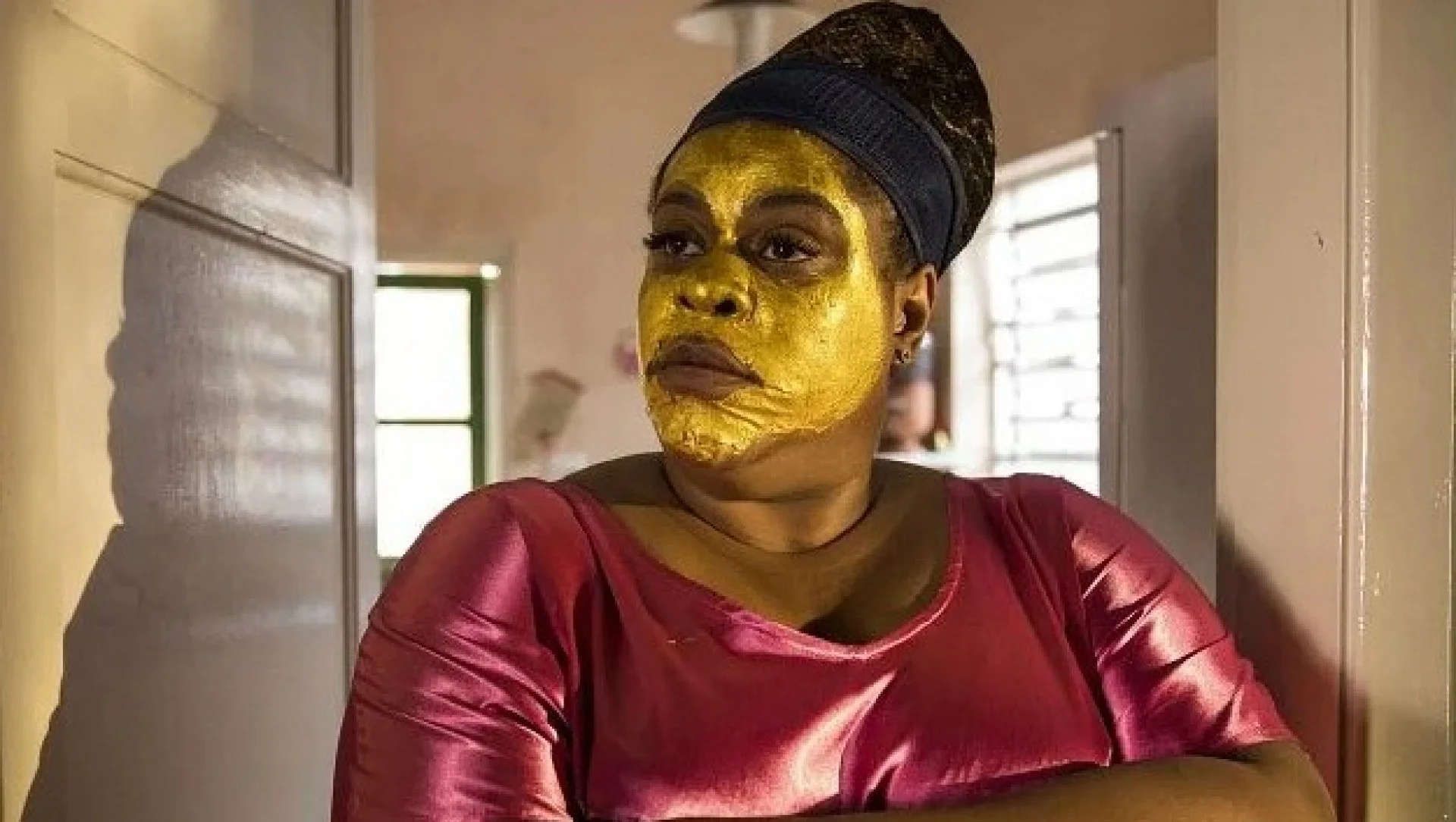 Mulher de braços cruzados com expressão séria, usando vestido rosa e máscara facial dourada.
